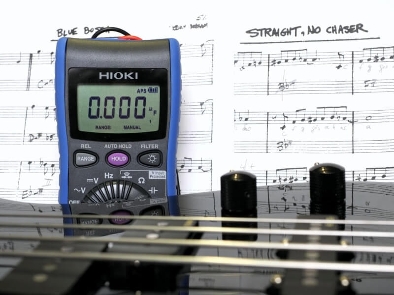 HIOKI digital multimeter in front of a bass guitar and sheet music during electric bass modding for timing and electrical measurement analysis.